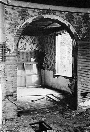 The arch separating the Dining Room and Parlor was originally draped with fabric. A plaster cove trims the ceiling above original wallpaper. A cast iron grate covered the floor opening in foreground, allowing heat to rise from the wood furnace below.