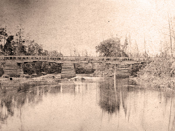 An early example. This bridge over the Whitemouth River in southeastern Manitoba was photographed in the mid-1870s by Winnipeg photographer Simon Duffin. Its design was typical for the day: wooden cribs supporting horizontal wooden spans, just wide enough for a single horse-drawn wagon to cross. Such bridges were frequent victims of shifting spring ice and flood water, and eventual decay of their unpreserved wood.