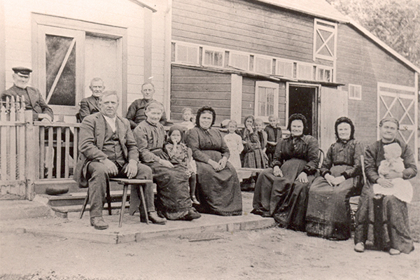 A group of Sommerfelder Mennonites visiting in Rosenbach, Manitoba, 1919. On the fenced porch is (L-R): Frank Enns, Isaac Wiens, and Rev. Wilhelm Friesen. Front row: Johann Braun, Maria (Bergmann) Braun, unidentified child, Katharina (Braun) Friesen, Katharina (Esau) Wiens, and Elizabeth (Esau) Enns. The woman on the far right and the children in the background are not identified.